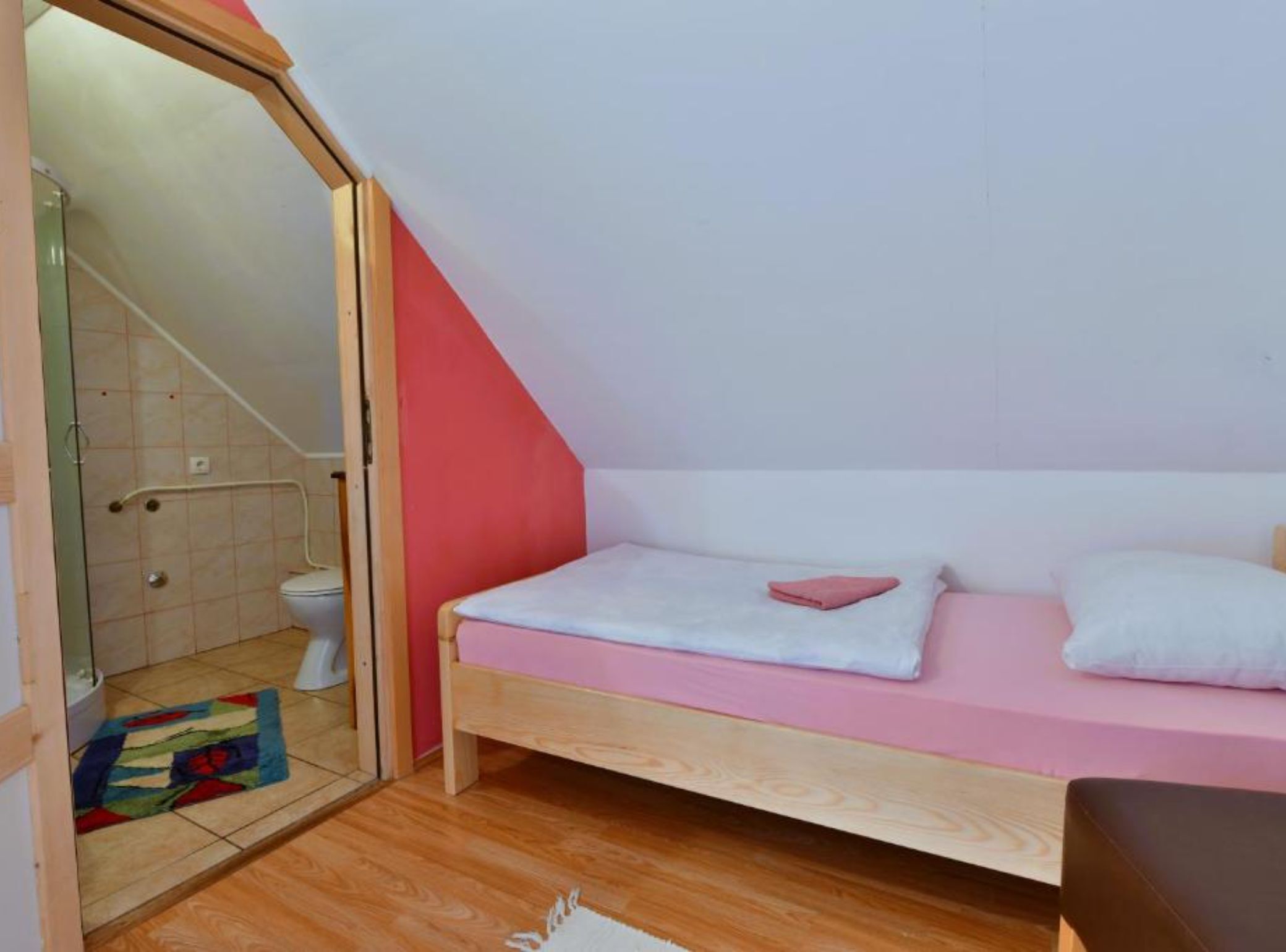 Un petit appartement d’une seule pièce, avec un plafond en pente, un lit en bois, une petite salle de bain avec des toilettes et une douche, et un tapis coloré sur le sol.