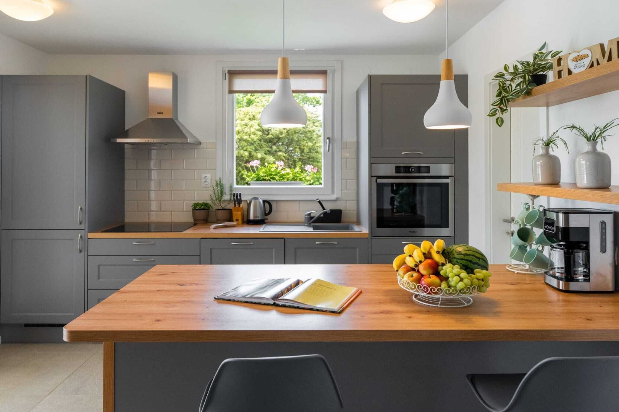 Ett modernt kök med grå skåp, en bänkskiva i trä, ett fönster med utsikt över grönområden, en kaffemaskin och en fruktskål.