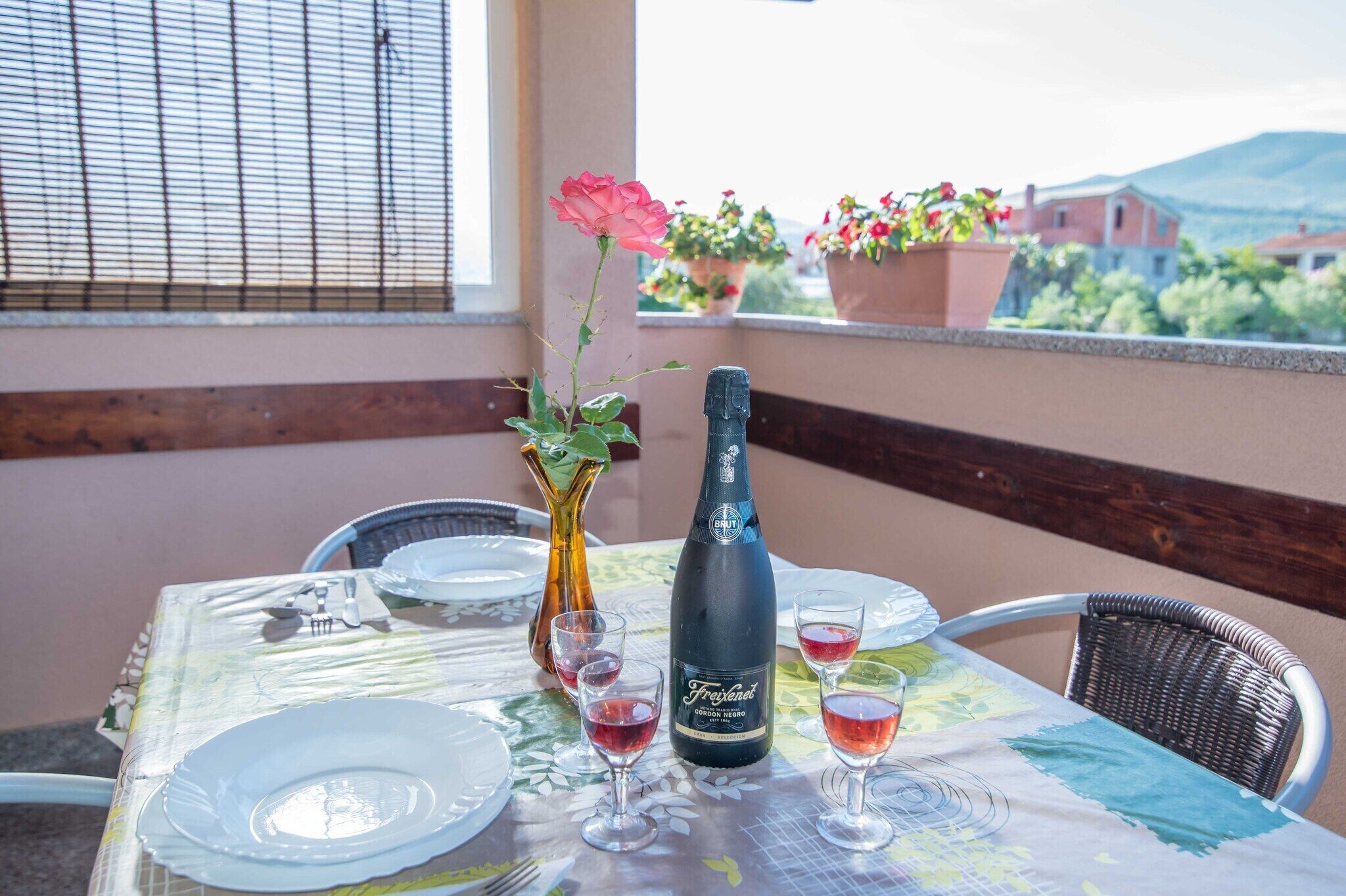A table set for a meal with a bottle of Freixenet champagne, a vase with a pink flower, and two glasses of red wine.