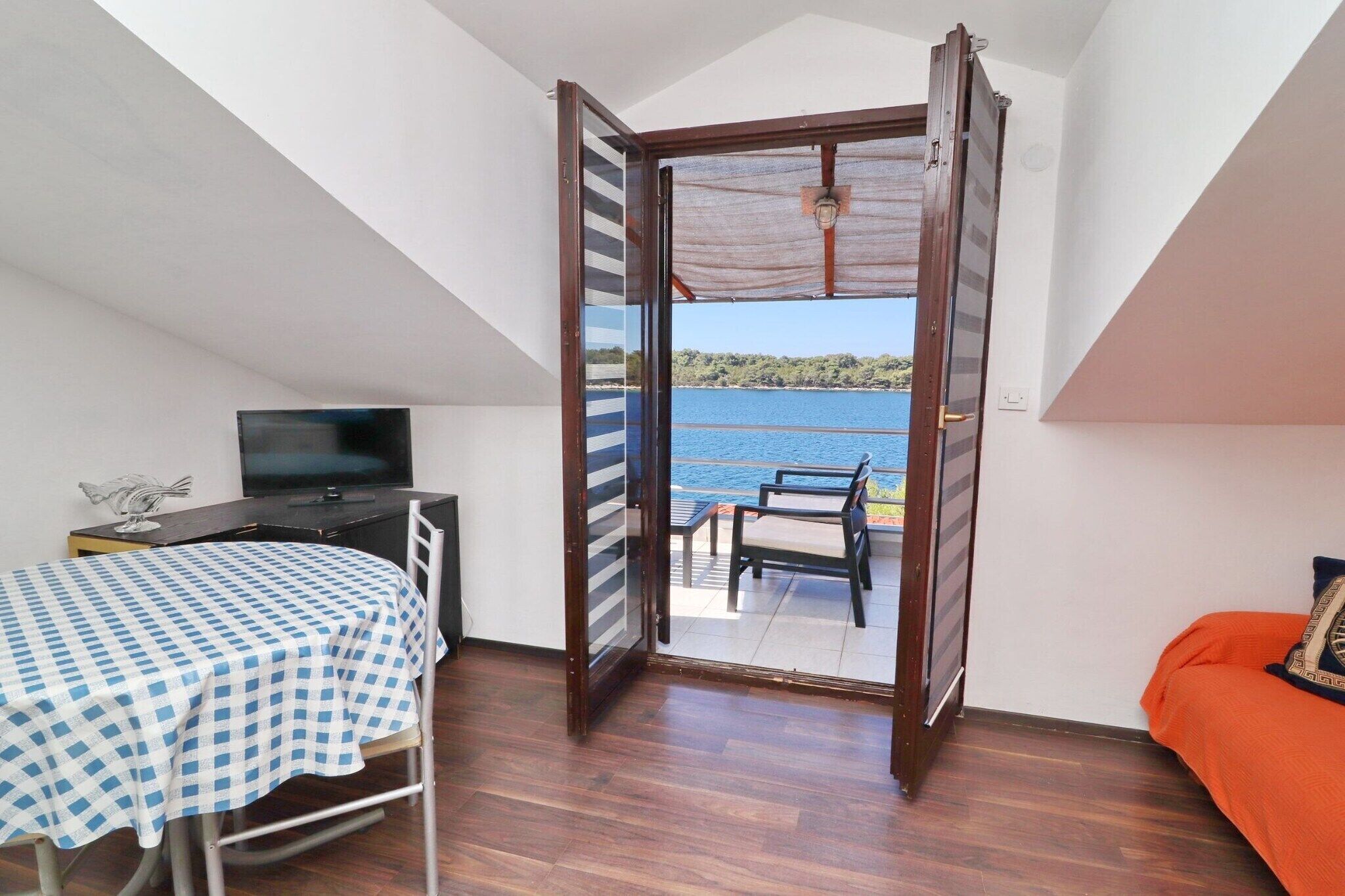 A room with a bed, a TV, a table with a checkered tablecloth, a balcony with a table and chairs, and a view of the sea.