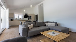 A modern living room with a grey sectional sofa, a wooden coffee table, and a kitchen in the background. - Three Bedroom Villa with Swiming Pool (Rasa)