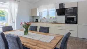 A modern kitchen with white cabinets, a wooden dining table with grey chairs, and a window with white curtains. - Three Bedroom Villa with Swiming Pool (Rasa)