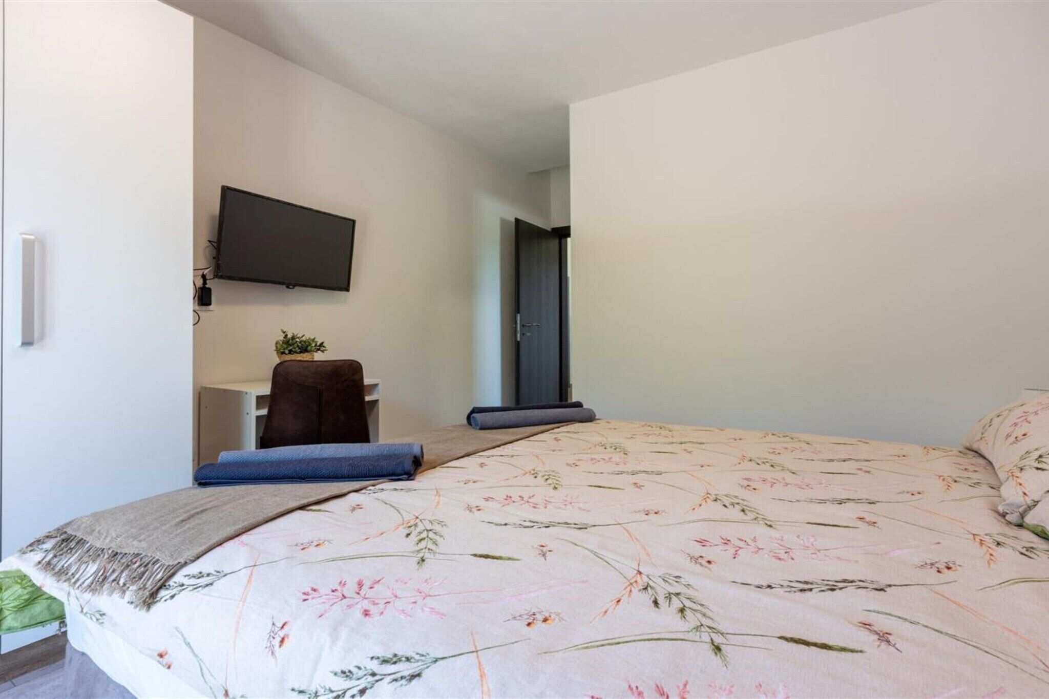 A bedroom with a bed, a flat-screen TV mounted on the wall, a desk with a chair, and a door leading to another room.