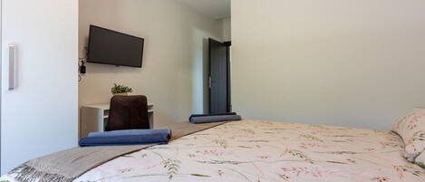 A bedroom with a bed, a flat-screen TV mounted on the wall, a desk with a chair, and a door leading to another room.
