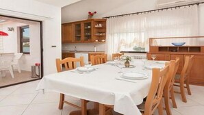 Un comedor con una mesa preparada para seis, sillas de madera, mantel blanco y una cocina con armarios y un ventanal con cortinas.