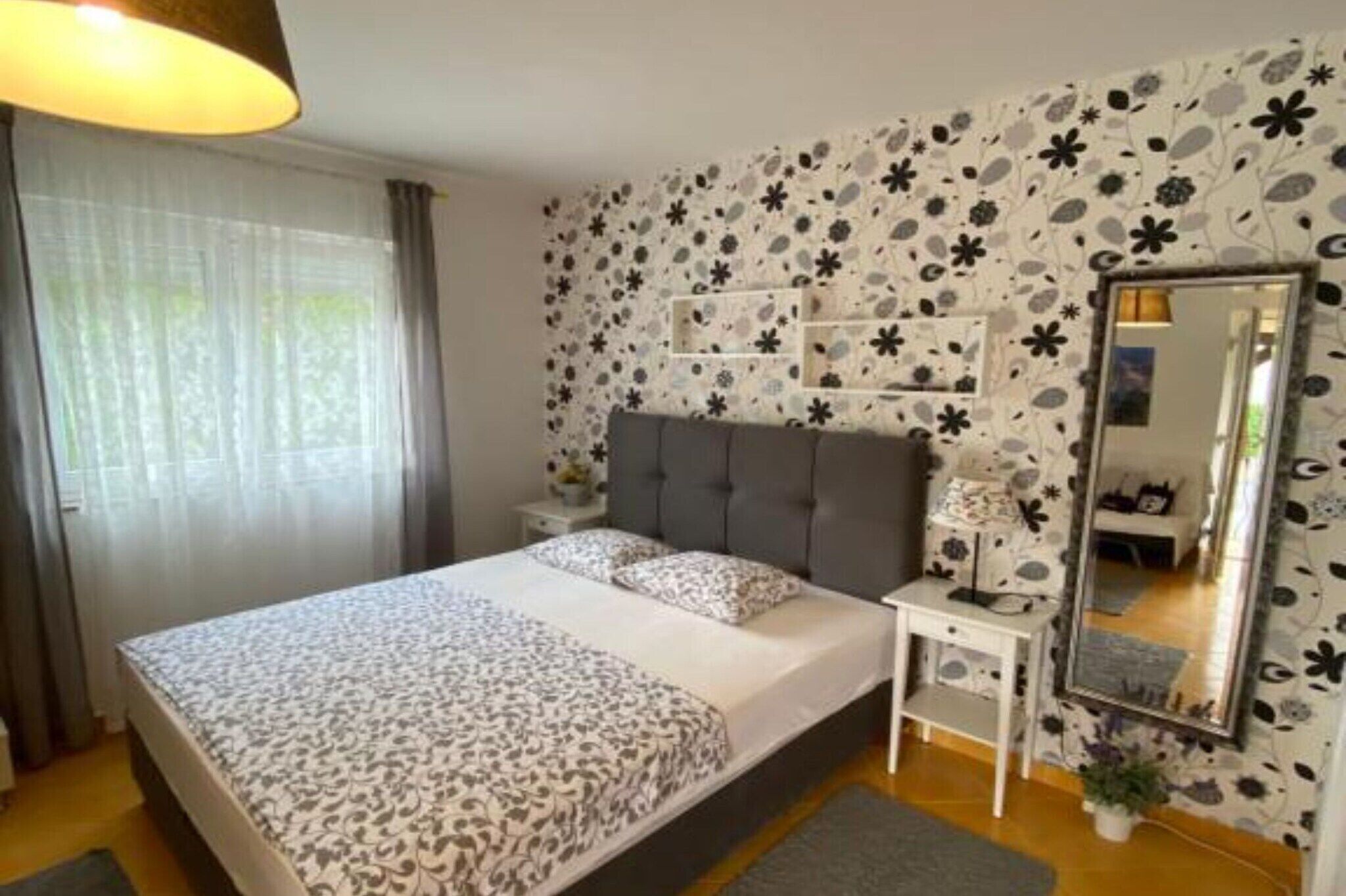 A bedroom with a bed, a bedside table, a mirror, and a wall with floral wallpaper.