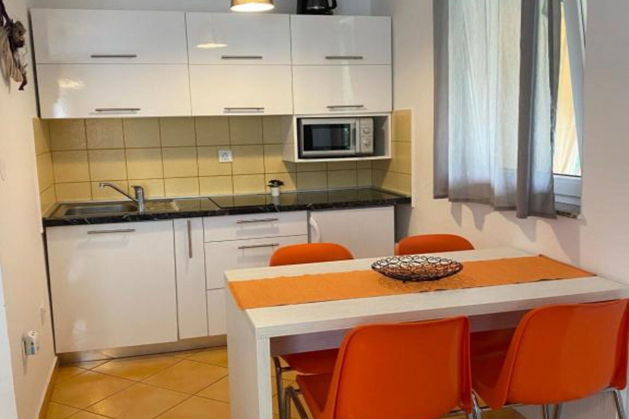 A compact kitchen with white cabinets, a microwave, and a sink. There is a dining table with orange chairs and a placemat.