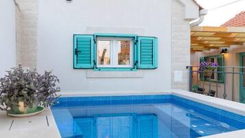Una piscina con vista a un edificio con contraventanas color turquesa y una planta en maceta.