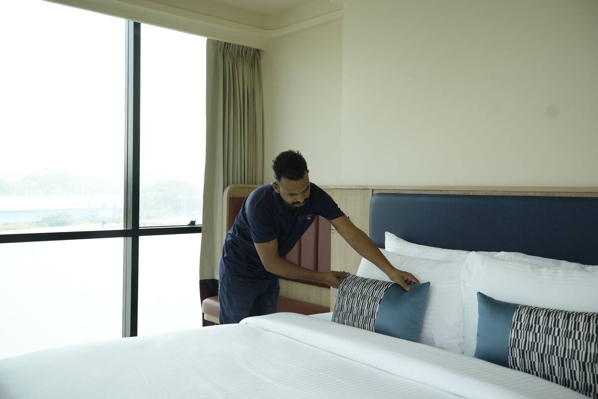A hotel room with a large bed, a view of the outdoors, and a person adjusting the bed linens.