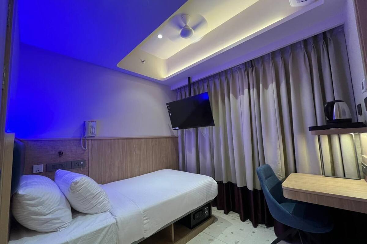 A compact hotel room with a single bed, a desk, a chair, a flat-screen TV mounted on the wall, and a ceiling fan.