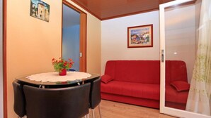 Una habitación con un sofá rojo, una mesa de comedor con sillas, una pequeña mesa roja con flores y una puerta que da a otra habitación.