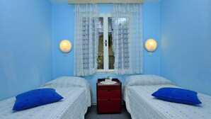 Una habitación con dos camas individuales, un pequeño aparador de madera y una ventana con cortinas de encaje.