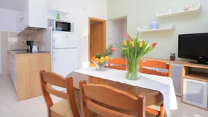 A compact kitchen with a microwave, refrigerator, and wooden cabinets. There is a dining table with chairs and a vase of flowers. A television is on a stand to the right.