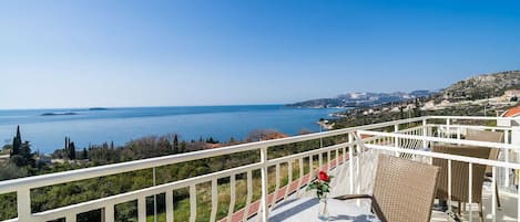 A balcony with wicker furniture, a small table with a vase, and a view of the sea and distant hills.
