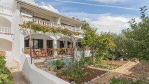 Een wit gebouw met een overdekte patio, zitplaatsen buiten en een tuin met verschillende planten en een tuinslang.