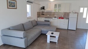 Ein kompakter Wohnraum mit grauem Ecksofa, einem kleinen weißen Tisch, einer Küchenzeile mit weißen Schränken, einem weißen Kühlschrank und einem Fenster, das für natürliches Licht sorgt.