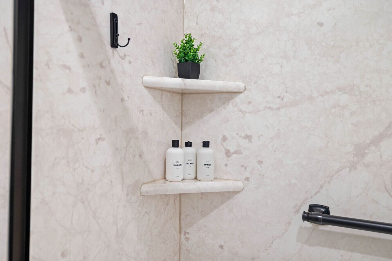 Ein Badezimmer mit Marmorwand, einem schwarzen Haken, einem kleinen Topfpflanzen und drei weißen Flaschen mit den Aufschriften „CONDITIONER“, „BODY WASH“ und „SHAMPOO“ auf einem Regal.