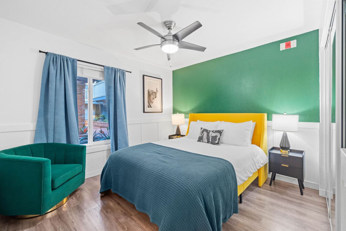 Una habitación de hotel con cama, un sillón verde, ventilador de techo y una ventana con cortinas.