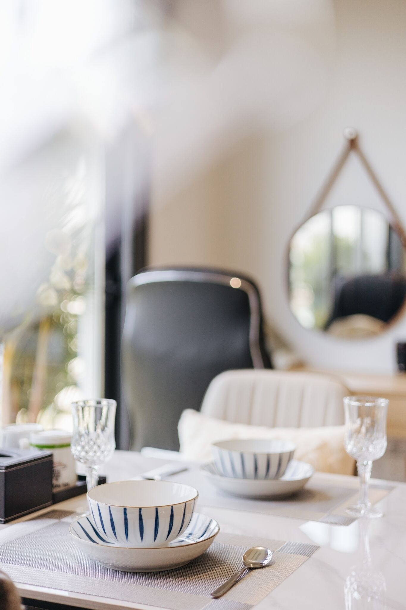 A dining table set with white and striped bowls, a spoon, and wine glasses. A black chair and a white chair are positioned behind the table.