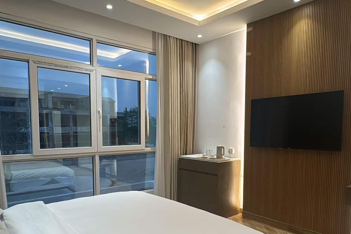 A modern hotel room with a large bed, a flat-screen TV mounted on the wall, and a window offering a view of the cityscape.