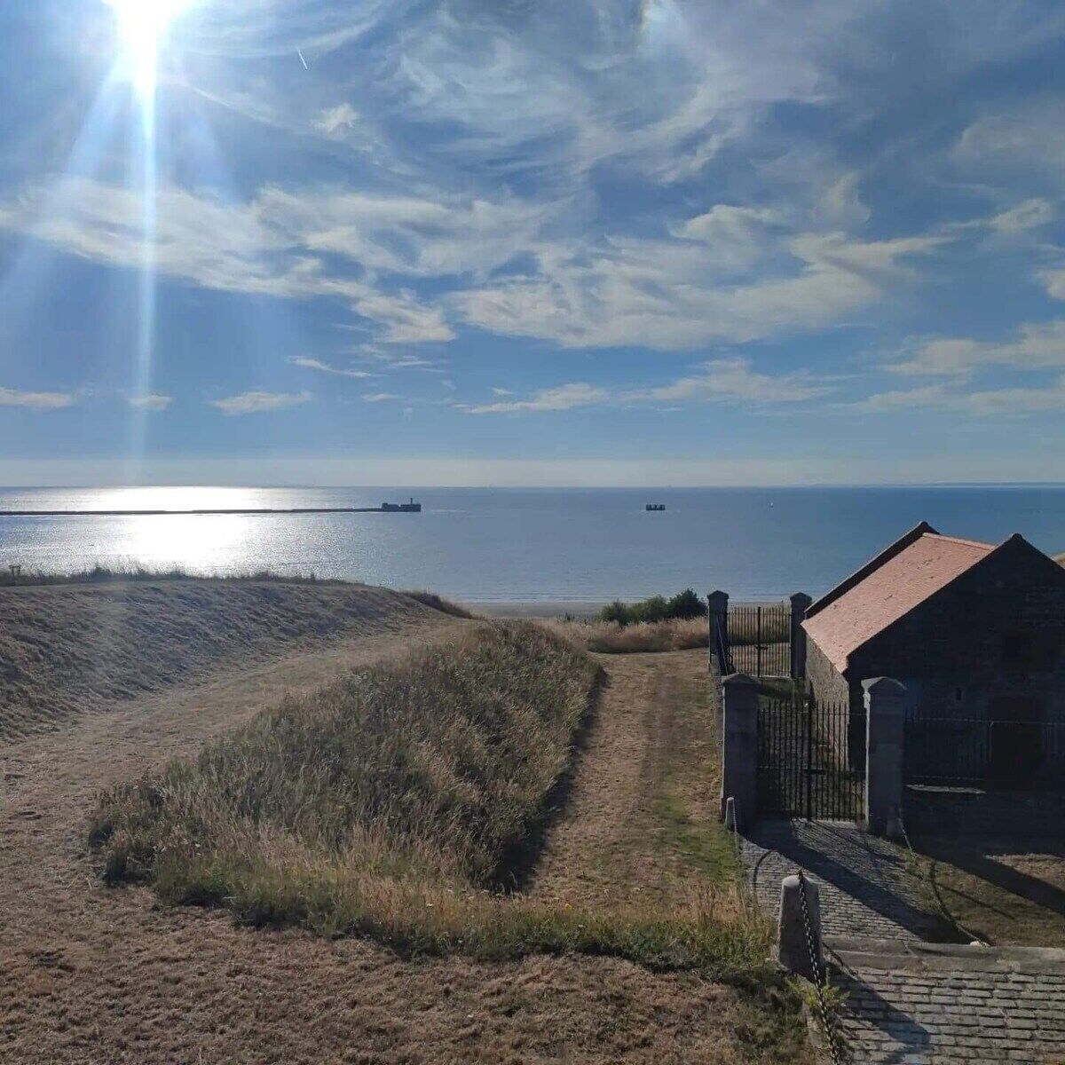 Ett kustlandskap med en sandstig som leder fram till en byggnad med ett rött tak.