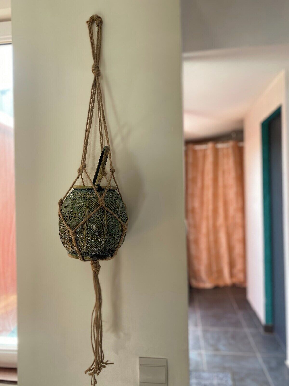 Une décoration murale en macramé, ornée d’un motif géométrique, est suspendue à un élément fixé au plafond dans un couloir. Le sol est carrelé et un rideau est visible en arrière-plan.