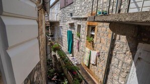Un estrecho callejón con edificios de piedra, un pequeño jardín con plantas en macetas, y un balcón visible con una silla.