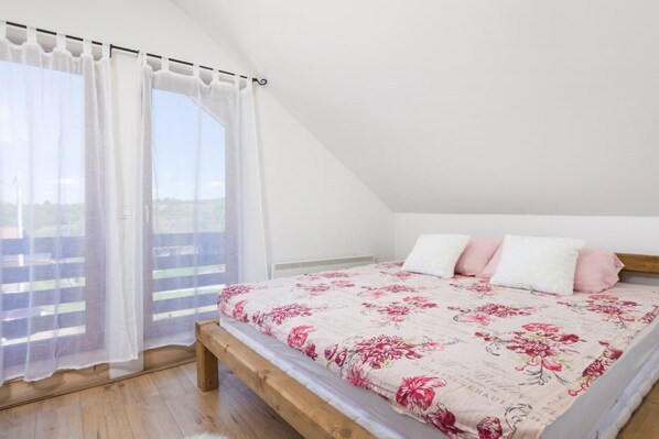 A bedroom with a wooden bed, white and pink bedding, and a view of a balcony with white curtains.