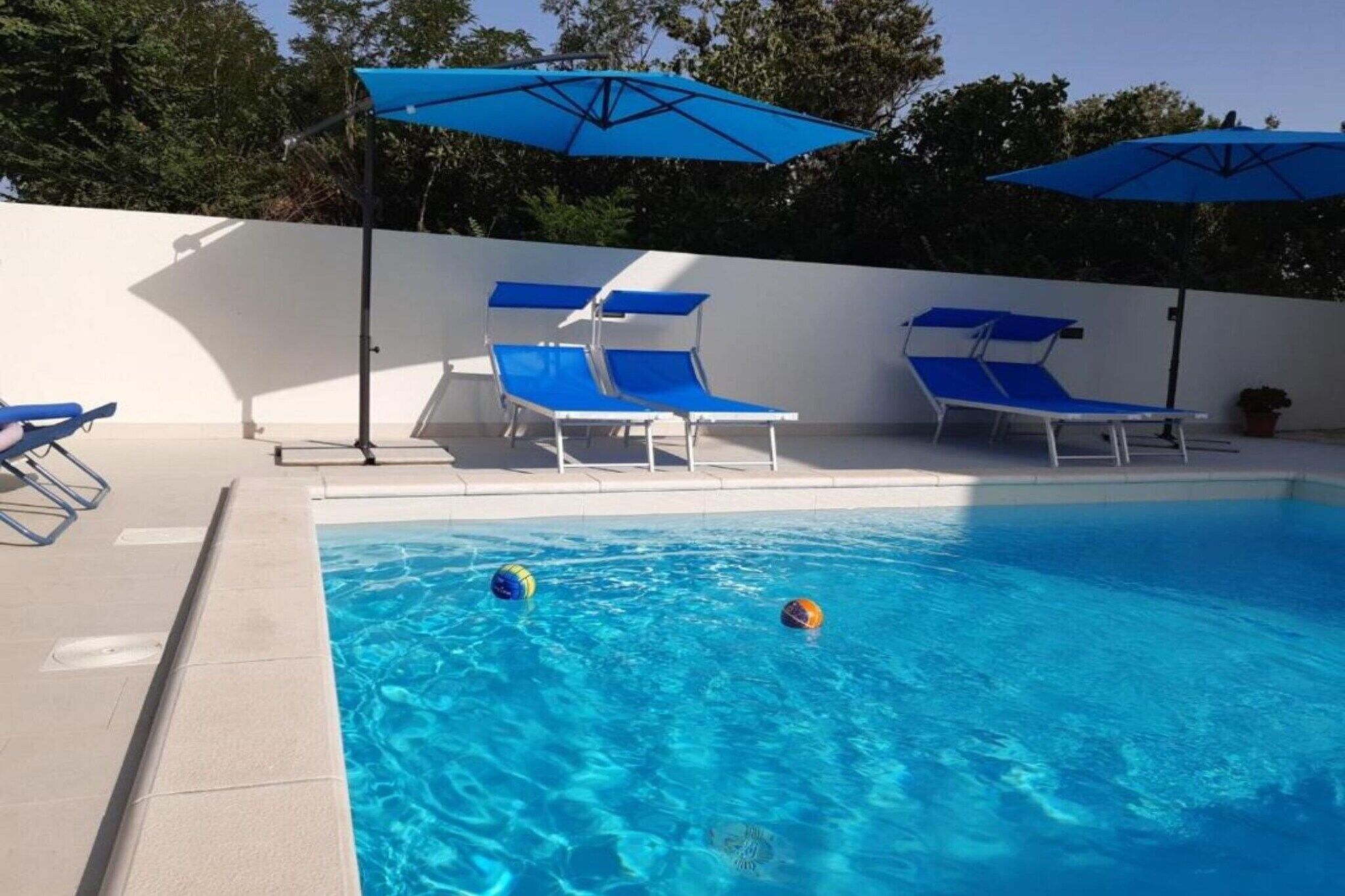 A swimming pool with blue lounge chairs and umbrellas, surrounded by a white wall and trees.