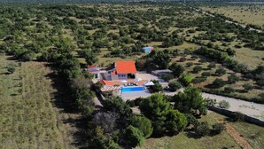 Une grande maison avec une piscine, entourée d’un paysage rural avec des arbres et des montagnes en arrière-plan.