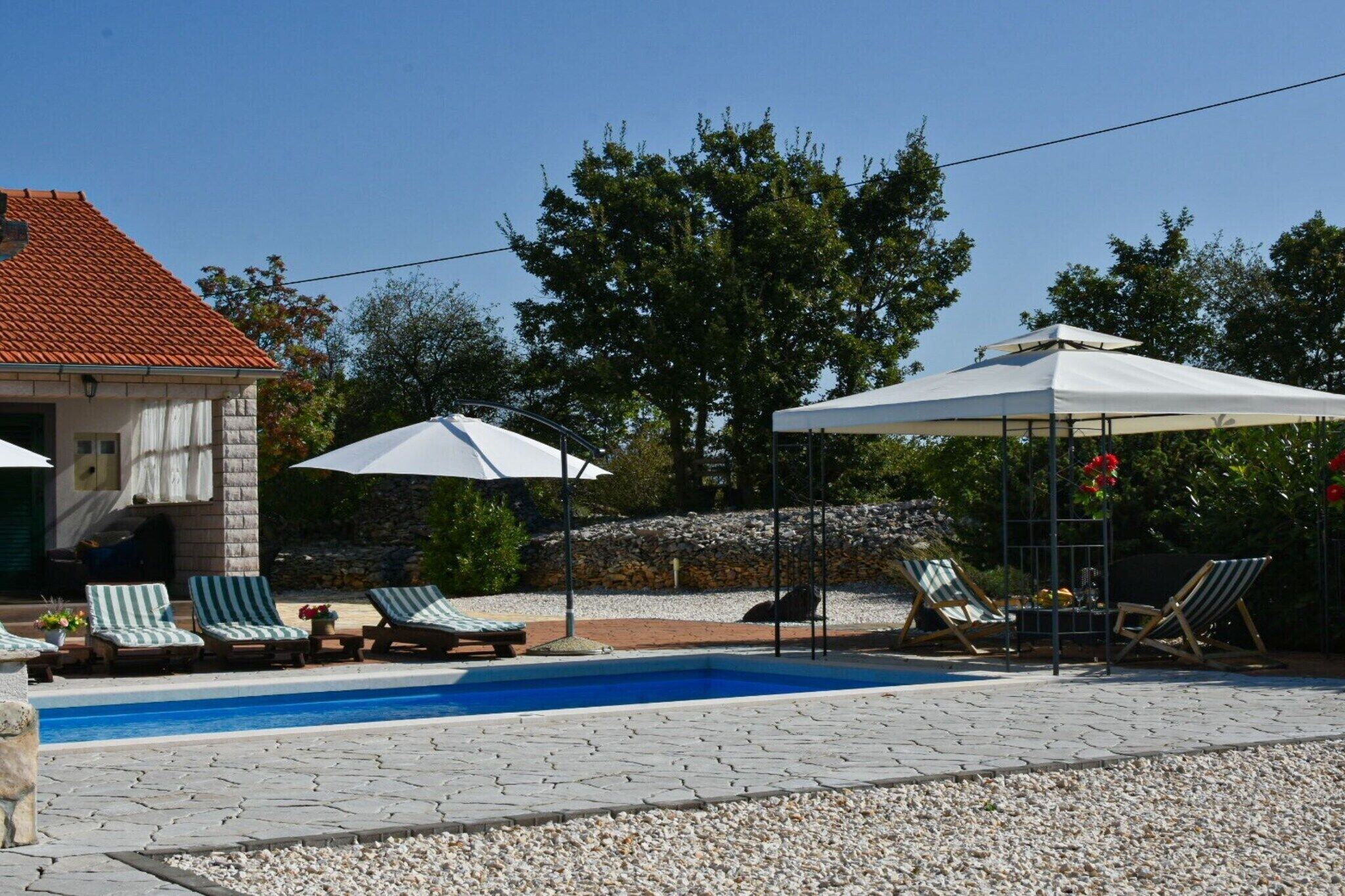 Un espace piscine aménagé avec des chaises longues, des parasols et un bâtiment en pierre doté d’un toit en tuiles.