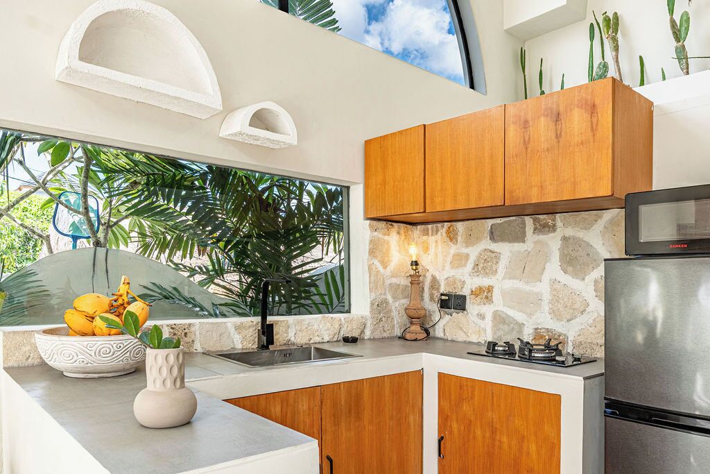 A modern kitchen with wooden cabinets, a stone backsplash, a stainless steel refrigerator, and a window with a view of greenery.