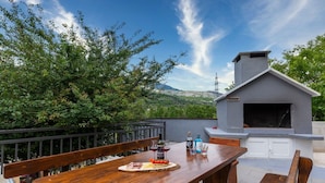 An outdoor dining area with a wooden table set for a meal, a built-in grill, and a view of a mountainous landscape. - Serene Rural Villa with Pool, Sun Deck, and Children's Play Area - Five Bedroom Villa (Tugare)