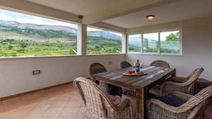 A dining area with a wooden table and wicker chairs, large windows offering a view of a green landscape and mountains. - Serene Rural Villa with Pool, Sun Deck, and Children's Play Area - Five Bedroom Villa (Tugare)