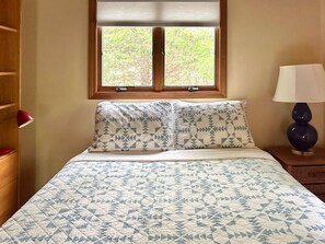 Een slaapkamer met een bed, twee kussens, een nachtkastje, een lamp, een raam en een houten garderobe.