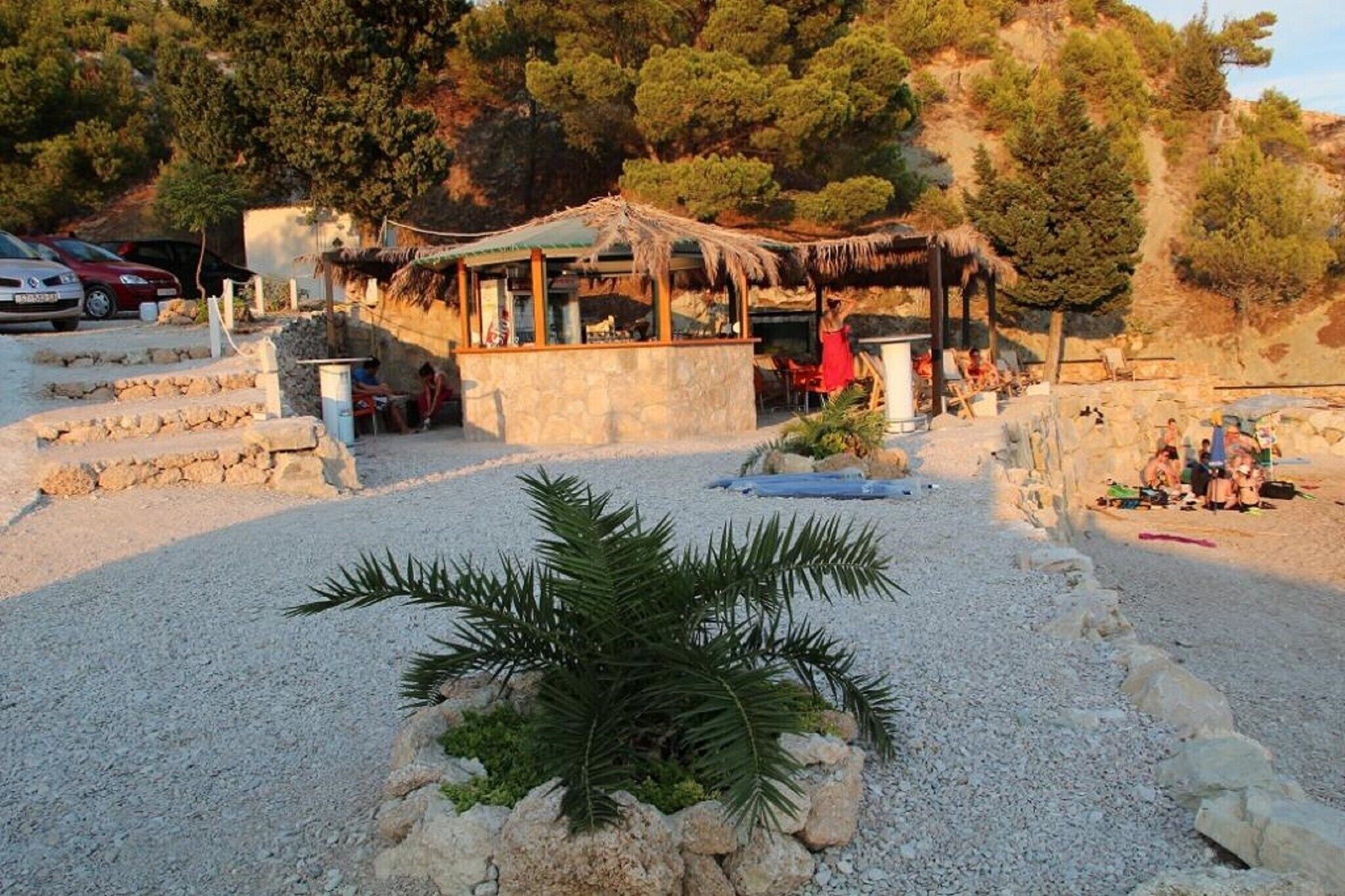 A rustic outdoor seating area with stone tables and chairs, surrounded by a gravel path and a palm tree.