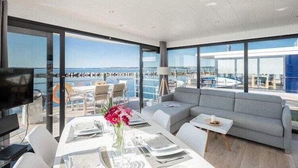 Ein modernes, helles Zimmer mit großem Fenster und Meerblick sowie Blick auf einen Poolbereich. Es gibt ein Sofa, einen Esstisch mit Stühlen und einen Fernseher.