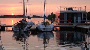Un port de plaisance avec des bateaux amarrés, un petit bâtiment doté d’un balcon, et un coucher de soleil en arrière-plan.