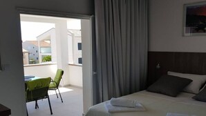 Habitación de hotel con cama, ventana con cortinas, balcón con sillas y vista a edificios.