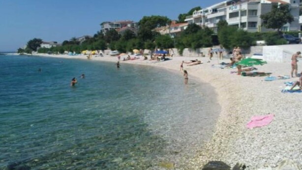 Una playa con aguas cristalinas, gente nadando y tomando el sol, y edificios al fondo.
