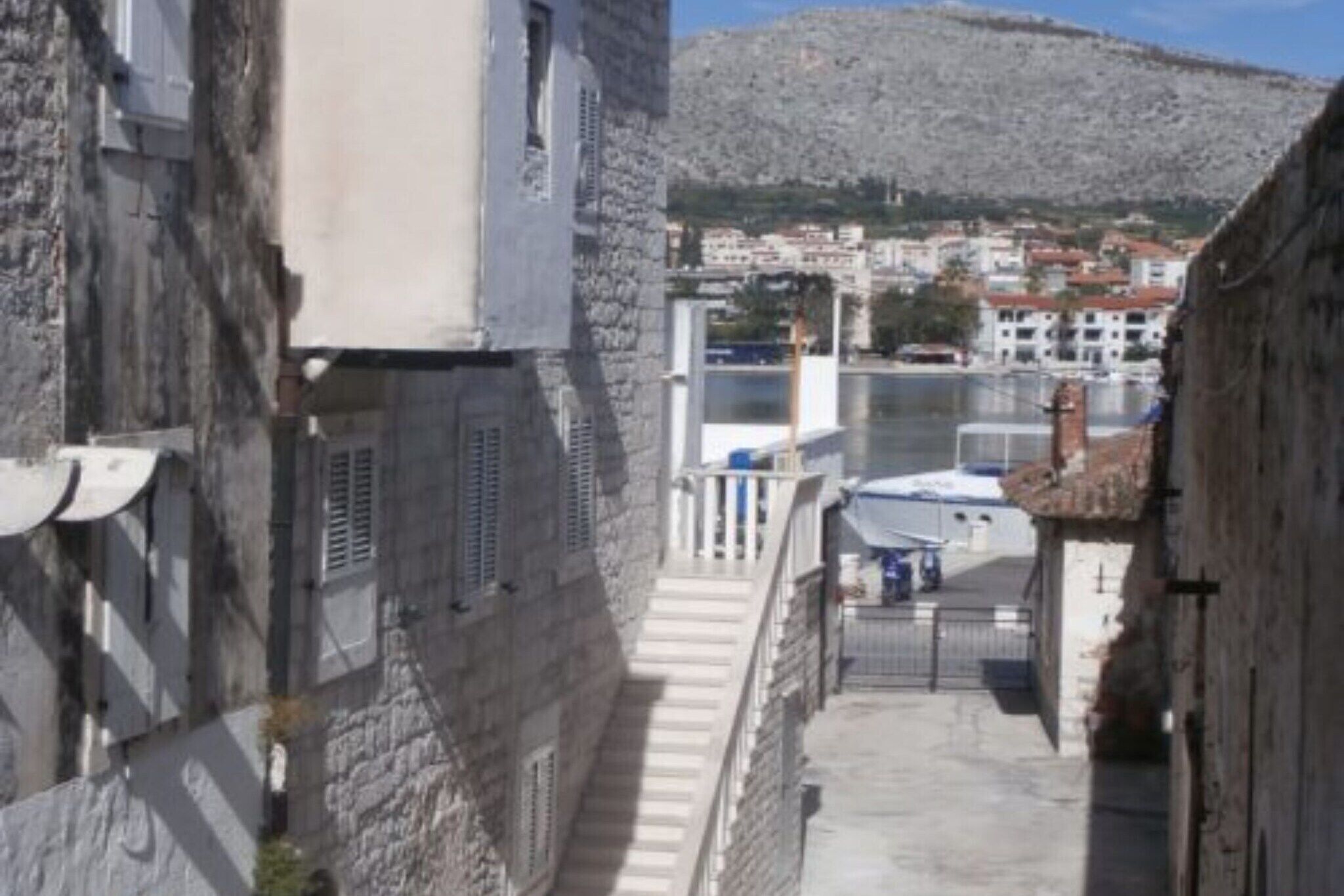 Ein schmaler Steinkorridor mit einer Treppe, die zu einem Tor führt, mit Blick auf ein Gewässer und eine an einem Hügel liegende Stadt.