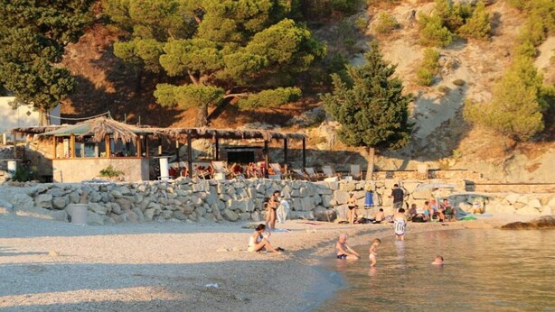 A beach with a stone wall, a thatched-roof structure, and people swimming and relaxing. - Apartments Tafra - One Bedroom Apartment (Lokva Rogoznica)