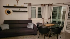 Una sala de estar con un sofá de color oscuro, una mesa de comedor con cubierta de vidrio y cuatro sillas, y un reloj montado en la pared.