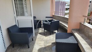 Un balcón con una mesa redonda y cuatro sillas, un pequeño rincón de descanso, y vistas a edificios al fondo.