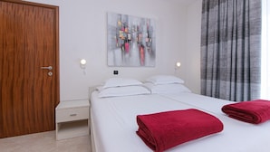 A hotel room with a wooden door, a bed with white linens, a red towel, a gray abstract painting, and a window with gray curtains.