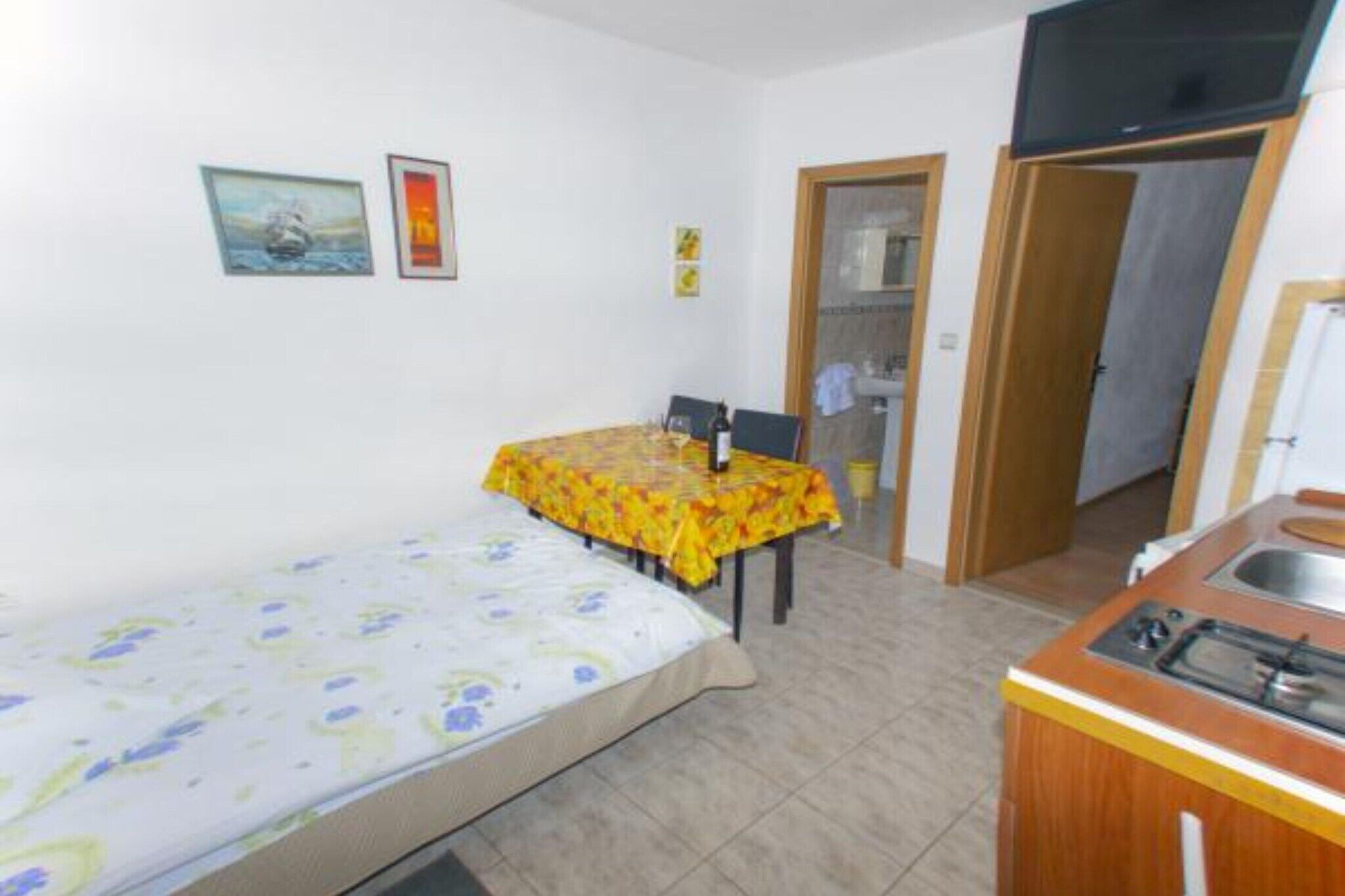 Una habitación pequeña y compacta con cama, una mesa con mantel amarillo, una kitchenette con fregadero y una puerta que da a otra habitación.