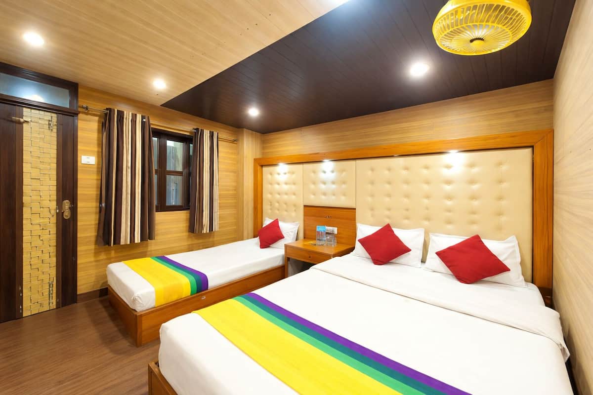 A hotel room with two beds, wooden flooring, a large window with curtains, a headboard with a light fixture, and a small table with a lamp.