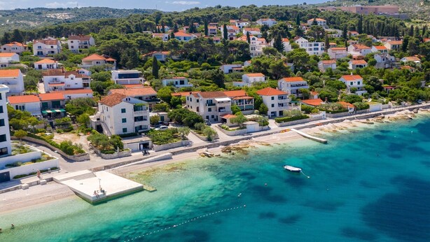 Luchtbeeld van een kustgebied met woningen, een steiger en helder turquoise water.