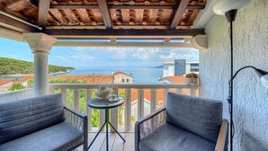 Een balkon met twee grijze stoelen, een kleine tafel met een vaas en een kopje, en een lamp. Het balkon heeft uitzicht op de kust met gebouwen en een heldere lucht.