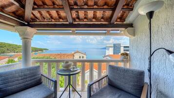 A balcony with two grey chairs, a small table with a vase and cup, and a lamp. The balcony overlooks a coastal view with buildings and a clear sky.